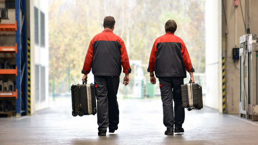 Zwei Mitarbeiter des mobilen Werkzeugbau-Services von weba in Arbeitskleidung, die mit Werkzeugkoffern in einer Werkshalle unterwegs sind. Sie sind auf dem Weg zur Installation oder Wartung von Umform- und Stanzwerkzeugen vor Ort beim Kunden.