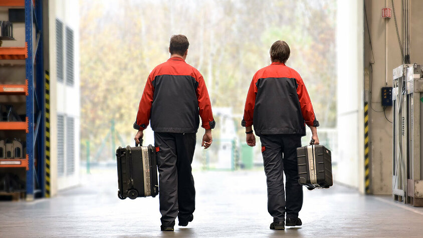 Zwei Mitarbeiter des mobilen Werkzeugbau-Services von weba in Arbeitskleidung, die mit Werkzeugkoffern in einer Werkshalle unterwegs sind. Sie sind auf dem Weg zur Installation oder Wartung von Umform- und Stanzwerkzeugen vor Ort beim Kunden.
