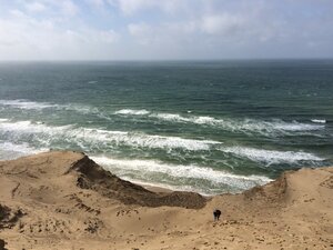 A person climbs a steep sand dune on the Danish coast, overlooking the rough sea and crashing waves under a cloudy sky.