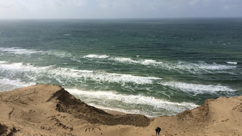 Person erklimmt eine steile Sanddüne an der dänischen Küste mit Blick auf die raue See und brechende Wellen unter bewölktem Himmel.