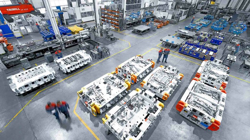 Modern production hall in tool manufacturing for stamping and pressing tools. A modern tool manufacturing facility with large stamping and pressing tools. The production hall is clean and well-organized, with several large tools placed on the floor. In the background, various machines and workbenches can be seen, while in the foreground, two employees in work uniforms are engaged in conversation. This image showcases the professional and efficient use of tools and machinery in the toolmaking industry, specifically for the automotive sector.