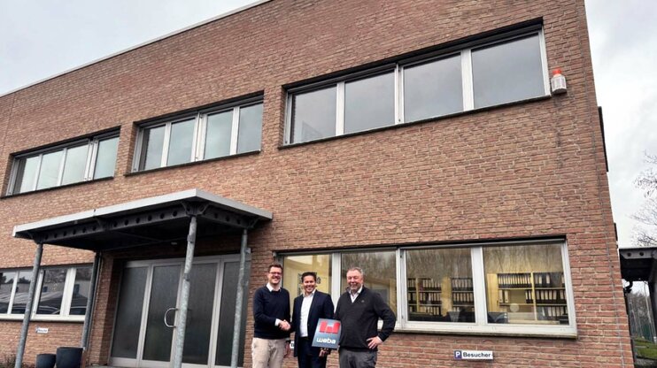 Strategische Expansion: weba stärkt Innovationskraft in Deutschland Drei Männer stehen vor einem Backsteingebäude, zwei schütteln sich die Hand, während der dritte ein Dokument mit weba-Branding hält.