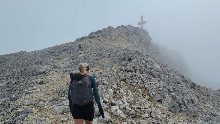 Bettina Gmainer mit Rucksack steigt einen felsigen Bergpfad hinauf, während sie sich einem Gipfelkreuz nähert. Der Himmel ist neblig, und die Atmosphäre wirkt rau und abenteuerlich.