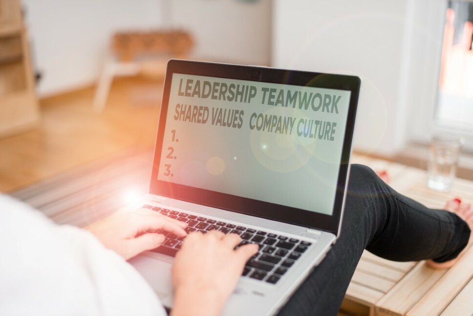 Eine Frau sitzt auf dem Boden mit einem Laptop auf ihrem Schoß. Auf dem Bildschirm sind die Wörter 'Leadership, Teamwork, Shared Values und Company Culture' zu lesen, während das Licht auf dem Laptop reflektiert wird. Das Bild symbolisiert wertegebundene Arbeitskultur bei weba für deine Karriere in Steyr.