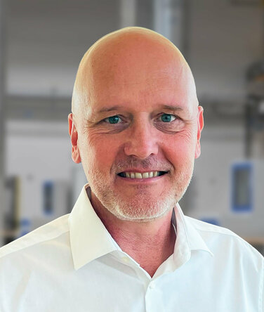 Porträt von Andreas Mehlmayer in einem modernen Industrieumfeld. Er trägt ein weißes Hemd und lächelt in die Kamera. Im unscharfen Hintergrund sind helle Fertigungseinrichtungen und Maschinen erkennbar – typisch für eine präzise und saubere Produktionsumgebung.