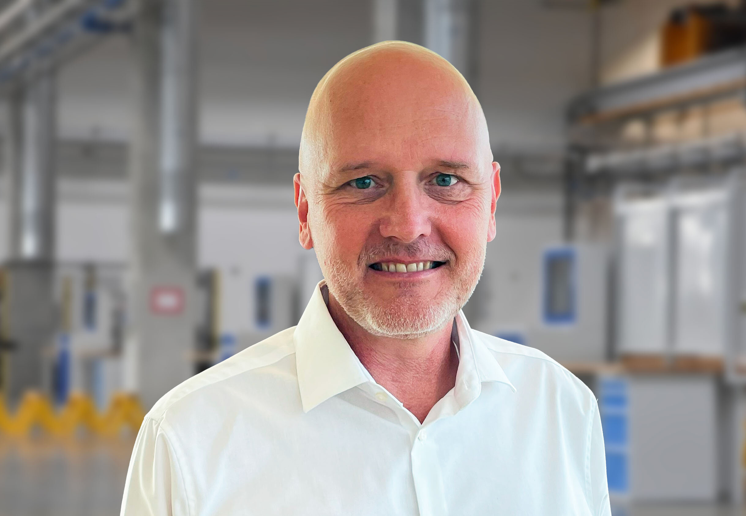 Porträt von Andreas Mehlmayer in einem modernen Industrieumfeld. Er trägt ein weißes Hemd und lächelt in die Kamera. Im unscharfen Hintergrund sind helle Fertigungseinrichtungen und Maschinen erkennbar – typisch für eine präzise und saubere Produktionsumgebung.