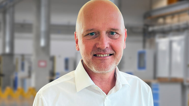Porträt von Andreas Mehlmayer in einem modernen Industrieumfeld. Er trägt ein weißes Hemd und lächelt in die Kamera. Im unscharfen Hintergrund sind helle Fertigungseinrichtungen und Maschinen erkennbar – typisch für eine präzise und saubere Produktionsumgebung.