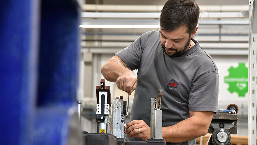 Ein Metalltechniker bei weba Werkzeugbau in Aktion, konzentriert auf den Zusammenbau von Messaufnahmen in der Produktionshalle. Der Techniker, in sauberer und praktischer Arbeitskleidung, arbeitet präzise an einem gut beleuchteten Arbeitstisch. Umgeben von verschiedenen Werkzeugen und Bauteilen, zeigt das Bild den Detailreichtum und die Präzision, die im Werkzeugbau erforderlich sind. Die Szene vermittelt ein Gefühl von Fachwissen und Engagement im Produktionsprozess, charakteristisch für die hohe Qualitätsstandards bei weba. Das Bild steht symbolisch für Jobs in Steyr für Berufserfahrene und Profis bei weba Werkzeugbau.