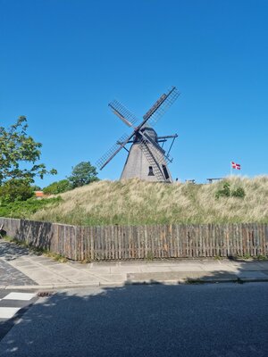 A historic windmill stands atop a dune-covered hill. Next to it, the Danish flag waves beneath a bright blue sky.
