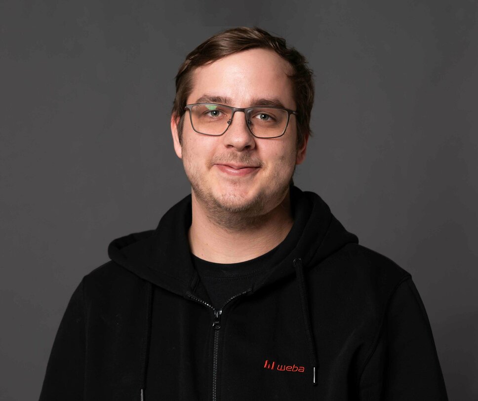 Porträtfoto des Lehrlingsausbilders bei weba Werkzeugbau für den Bereich Metalltechnik. Er lächelt in die Kamera und steht vor einem neutralen grauen Hintergrund.