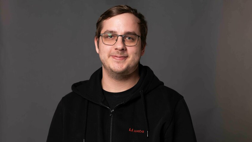 Porträtfoto des Lehrlingsausbilders bei weba Werkzeugbau für den Bereich Metalltechnik. Er lächelt in die Kamera und steht vor einem neutralen grauen Hintergrund.