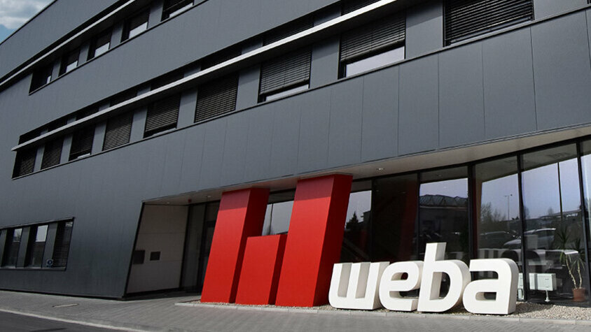 Foto der Front des weba-Gebäudes in Olomouc, gekennzeichnet durch eine anthrazitfarbene Fassade. Im Vordergrund steht ein auffälliges 3D-Logo in Rot und Weiß, das die Markenidentität von weba stark hervorhebt und einen markanten Kontrast zur dunklen Gebäudefarbe bildet.