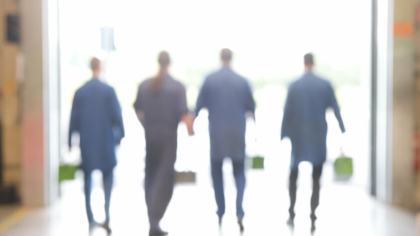 Vier Werkzeugbautechniker mit Werkzeugkoffern in der Hand, von hinten fotografiert, gehen durch ein großes Produktionstor heraus zum Montageeinsatz.