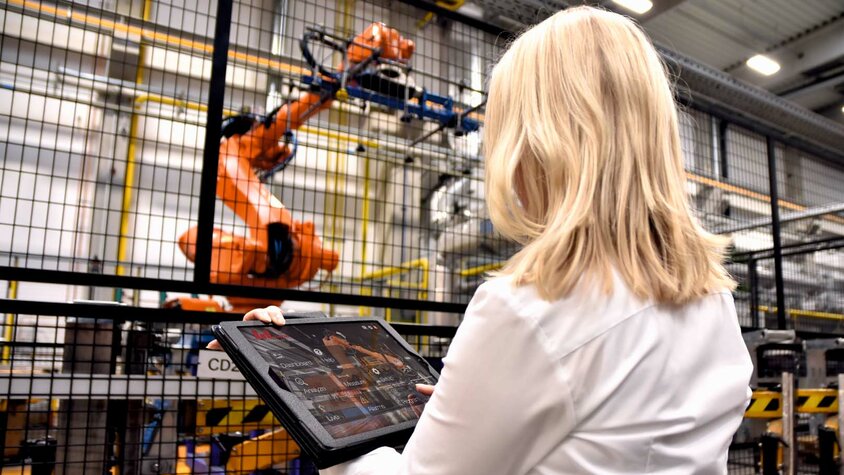 Eine Frau steht in einer industriellen Umgebung bei weba Werkzeugbau vor einem großen Robotergreifer einer Warmumformlinie. In ihren Händen hält sie ein Tablet, auf dem sie Daten überwacht oder Steuerbefehle für den Roboter eingibt. Der Fokus liegt auf der Interaktion zwischen moderner Technologie und menschlichem Eingreifen.
