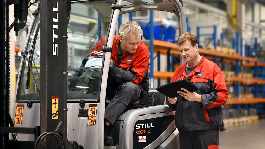 Zwei Werkzeugbautechniker besprechen die Logistik in der Serienproduktion für das Presshärten. Einer sitzt im Stapler und betrachtet das Tablet, das der andere Werkzeugbautechniker in der Hand hält.