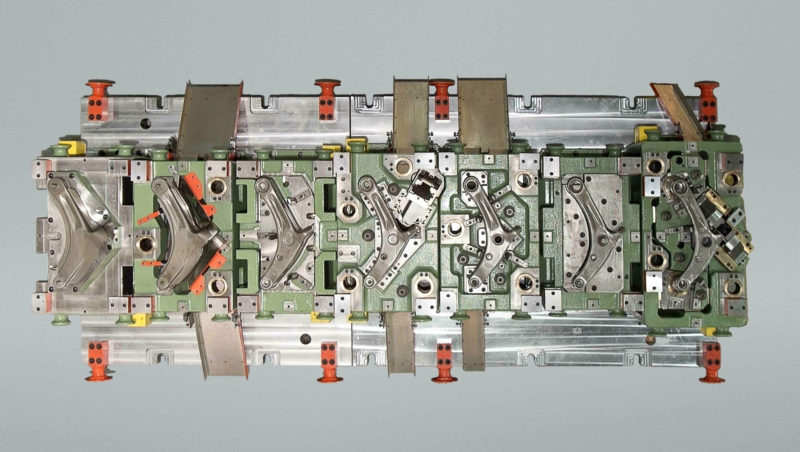 Top-Ansicht eines Transferwerkzeugs zur Herstellung eines Querlenkerarms; erkennbar sind mehrere Stationen zur Umformung und Positionierung des Bauteils in einem mehrstufigen Werkzeugaufbau.