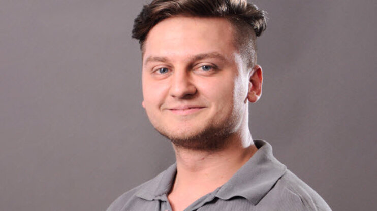 Porträt eines jungen Mannes mit kurzem, seitlich rasiertem Haar und einem freundlichen Lächeln. Er trägt ein graues Polohemd und blickt direkt in die Kamera. Der Hintergrund ist neutral und grau.