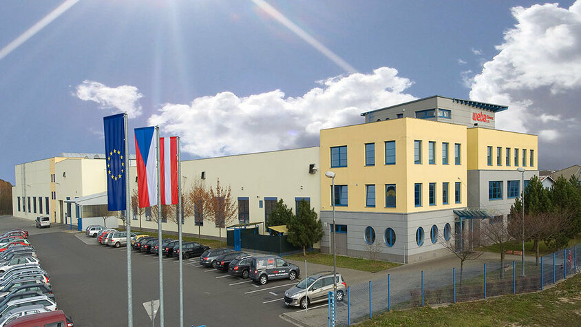 Außenaufnahme des Produktionsstandorts weba Olomouc, bei Sonnenschein unter einem blauen, fast wolkenlosen Himmel, vor dem Gebäude wehen Fahnen.