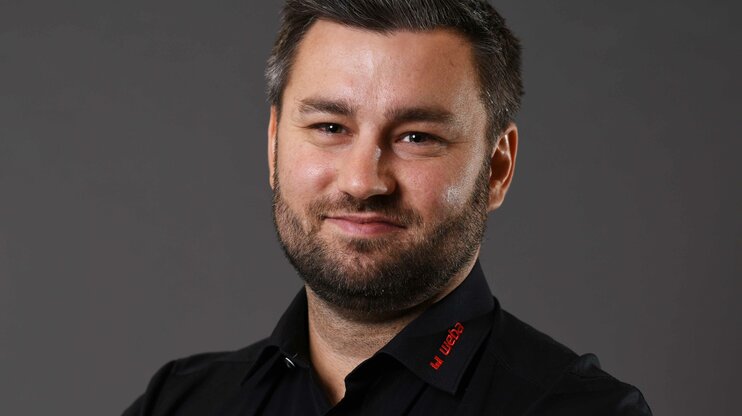 Alexander präsentiert sich freundlich und professionell: schwarzes Hemd mit rotem weba-Logo, vor grauem Hintergrund, direkt in die Kamera lächelnd.