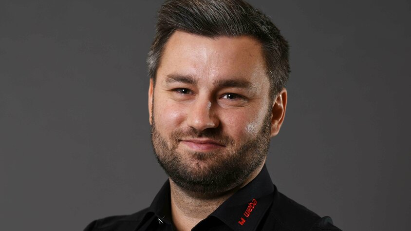 Alexander präsentiert sich freundlich und professionell: schwarzes Hemd mit rotem weba-Logo, vor grauem Hintergrund, direkt in die Kamera lächelnd.