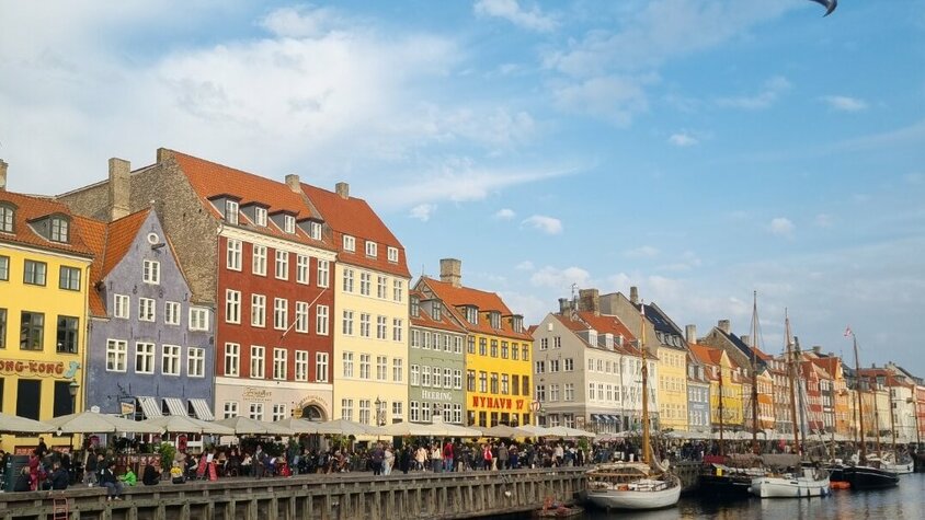 Nyhavn in Kopenhagen mit bunten Häusern, Booten im Wasser und vielen Menschen entlang der Promenade bei blauem Himmel.