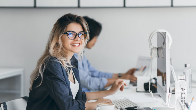 Junge Frau mit Brille sitzt lächelnd im modernen Büro am Schreibtisch, arbeitet am Computer, im Hintergrund Kollegen.
