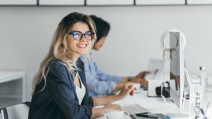 Junge Frau mit Brille sitzt lächelnd im modernen Büro am Schreibtisch, arbeitet am Computer, im Hintergrund Kollegen.