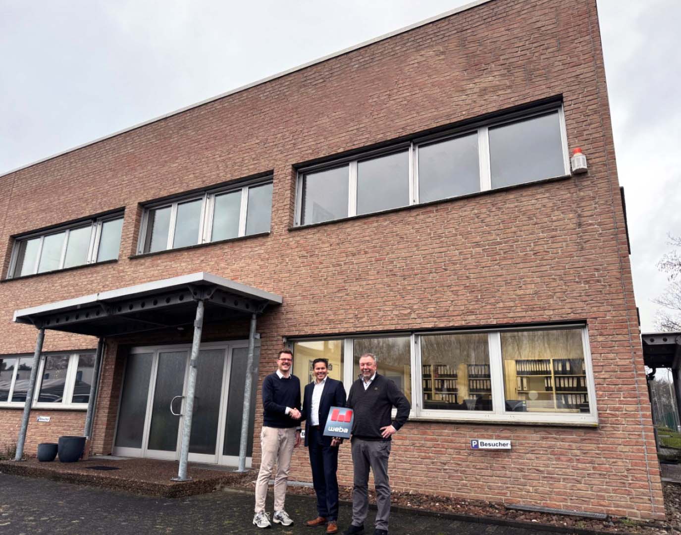 Drei Männer stehen vor einem Backsteingebäude, zwei schütteln sich die Hand, während der dritte ein Dokument mit weba-Branding hält.
