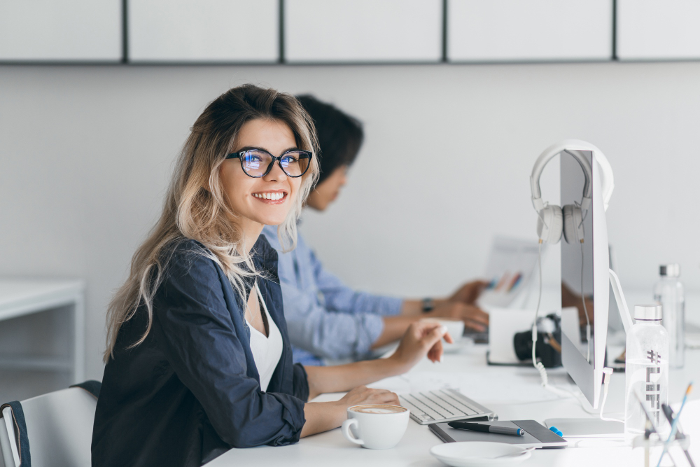 Junge Frau mit Brille sitzt lächelnd im modernen Büro am Schreibtisch, arbeitet am Computer, im Hintergrund Kollegen.